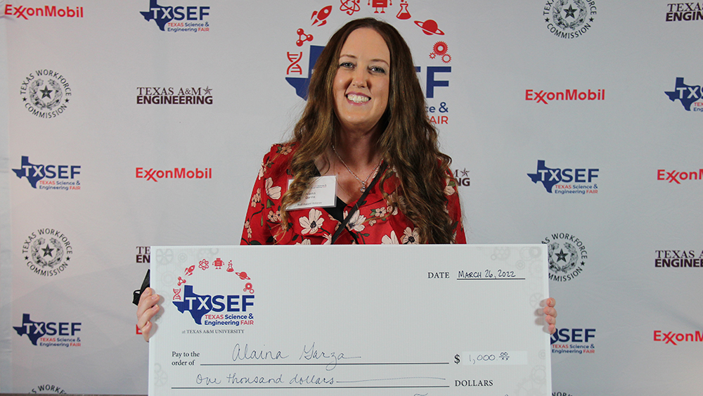 Alaina Garza holds a large white check against a backdrop with Texas Science and Engineering Fair logos