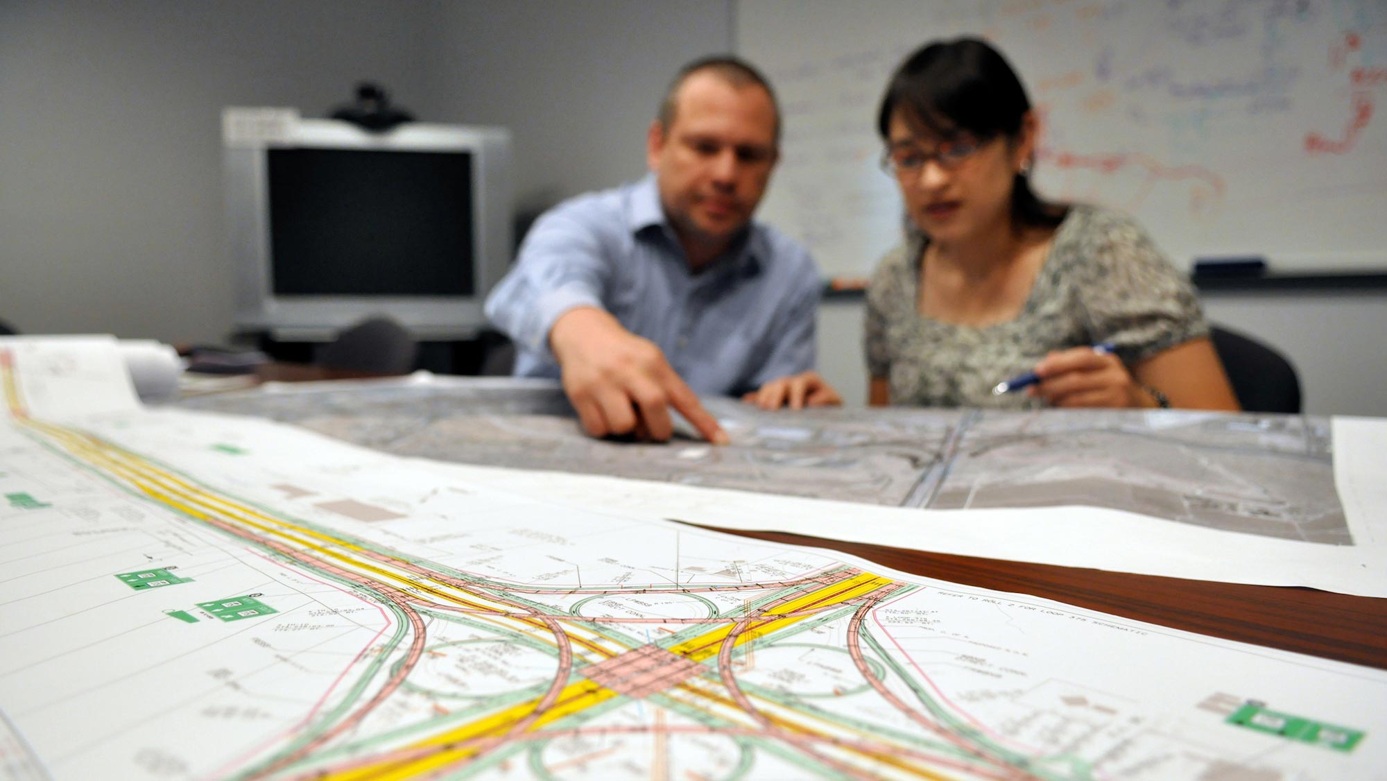 A woman with short black hair and glasses looks on as a man with a long-sleeved, blue button-down shirt points to something on the maps splayed across the table before them.