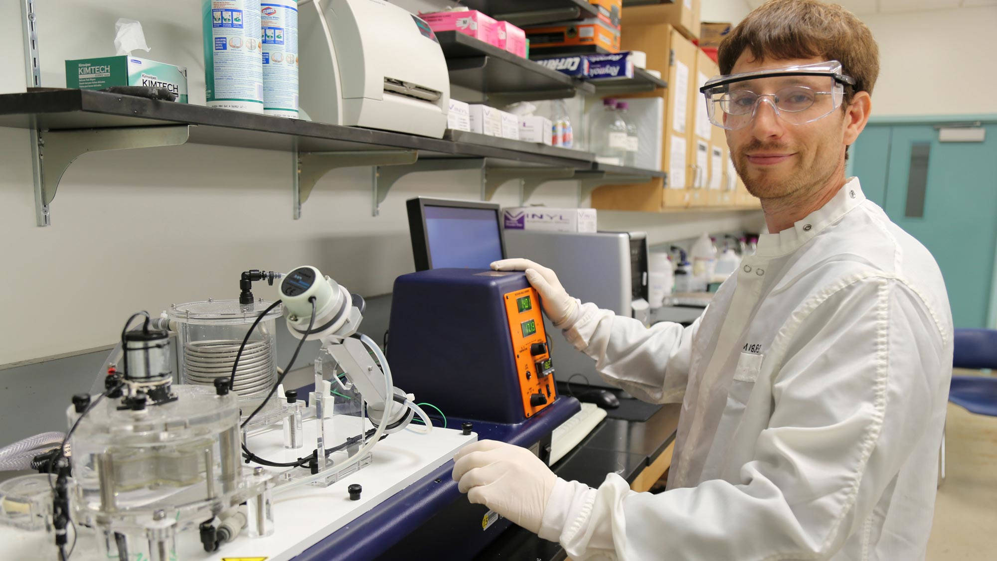 A smiling researcher with brown hair, wearing goggles over glasses, a white lab coat, and white latex gloves works with equipment in a chemical engineering laboratory setting.