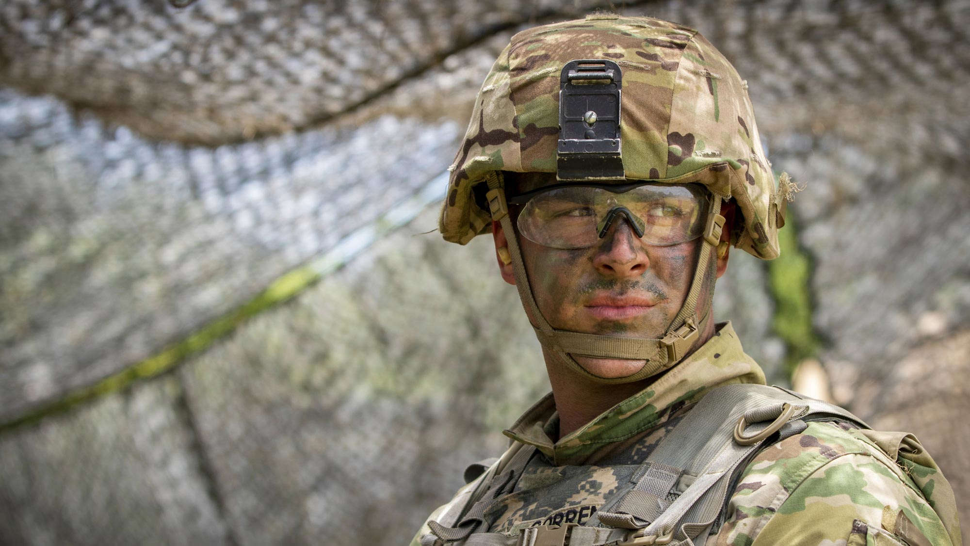An army soldier wearing military safety gear, helmet, and goggles, with his face covered in camouflaging paint.