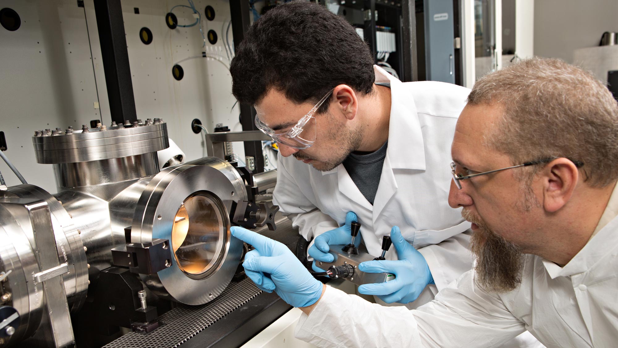 A male researcher with dark hair and goggles and an older male researcher with graying hair and a long beard and glasses observe equipment in a lab.