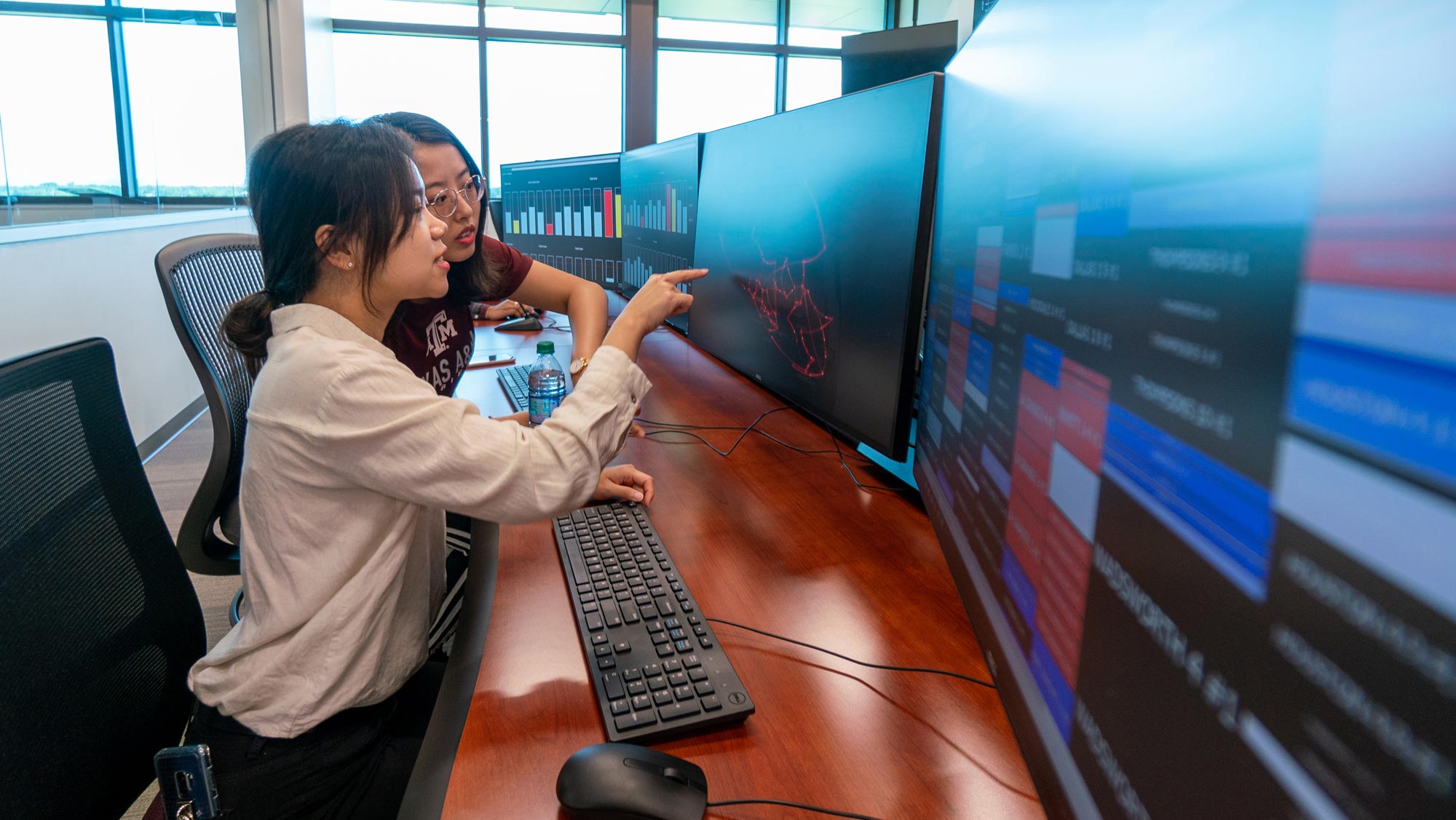 A female researcher with black hair pulled into a bun points to a computer monitor as a second female researcher with long black hair and glasses looks on. 