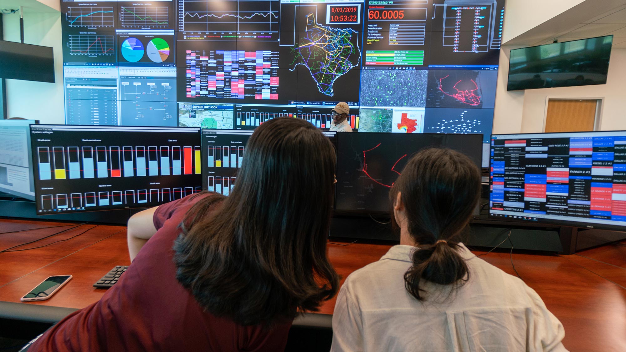 Two female researchers, one with long black hair and the other with black hair pulled into a bun, sit beside each other, with their backs to the camera, as they view a computer monitor. Behind the monitor is an entire wall of Smart Grid monitors.