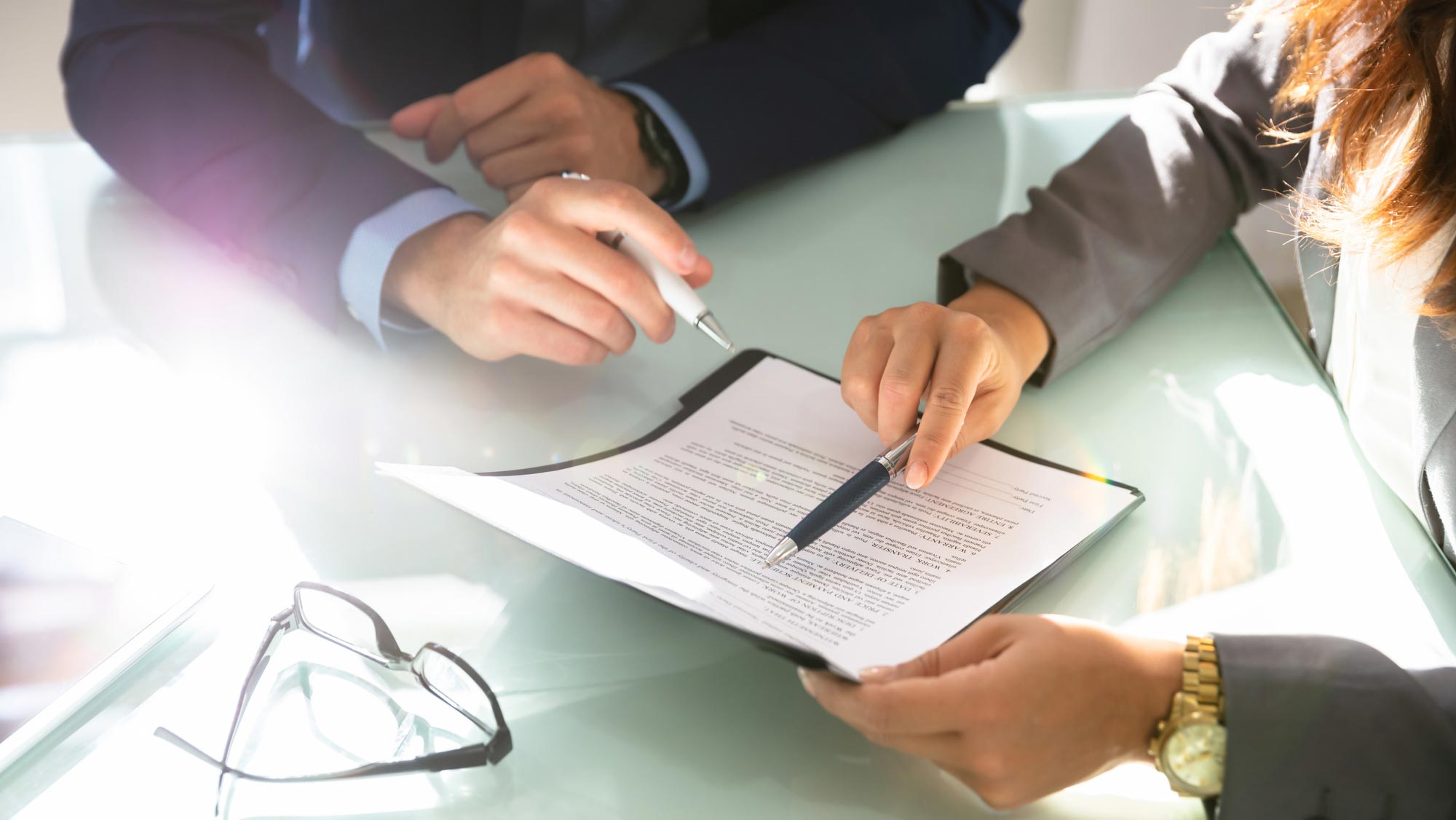 Two people's hands hold pens as they appear to review a document at a table. Someone's glasses also rest on the table.