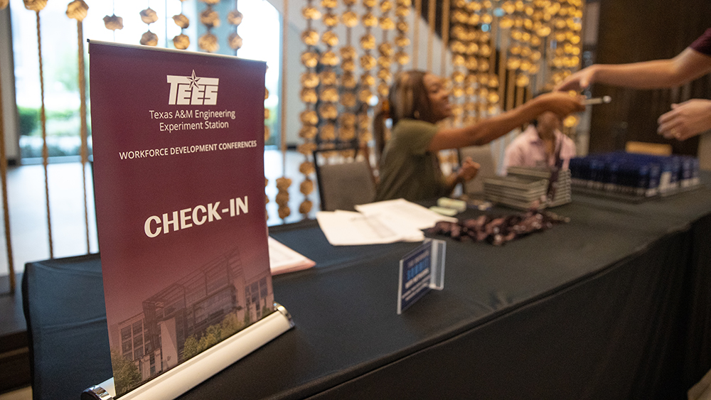 In the foreground, sits a small table-top maroon banner with the words TEES Texas A&amp;M Engineering Experiment Station Workforce Development Conferences and Check-in. In the background, two women sit at a table handing nametags to attendees. 