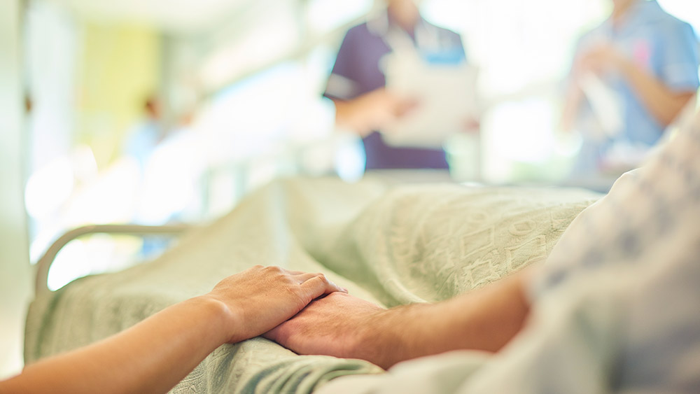 A visitor's hand holds a patient's hand in the bed of a hospital room. In the blurred background, two nurses are chatting about the patient's care.