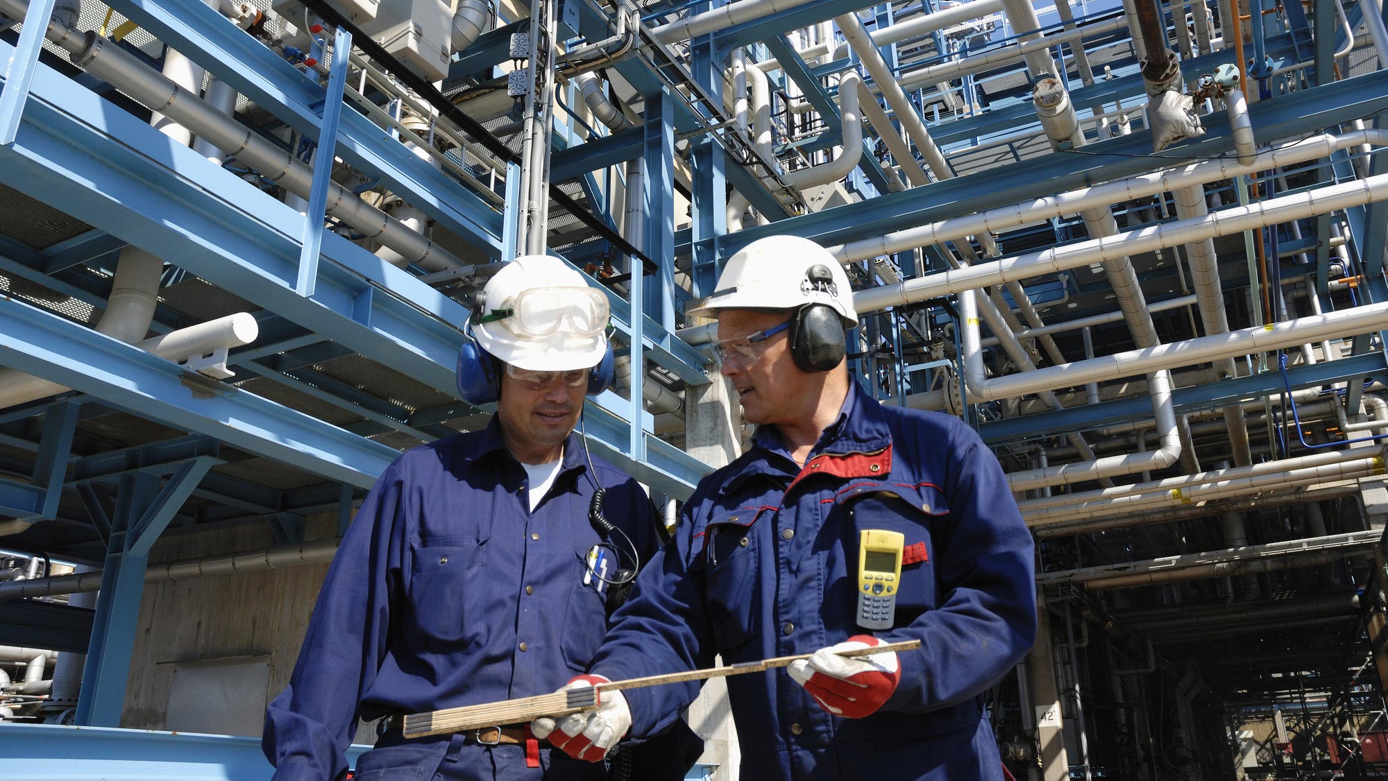 Two men wearing white construction hats, noise-canceling earmuffs, and blue jumpsuit's examine a ruler as they stand before an oil refinery.