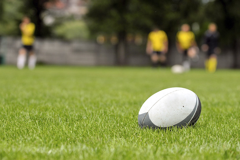 Gray rugby football on grass in foreground with people out of focus in the background