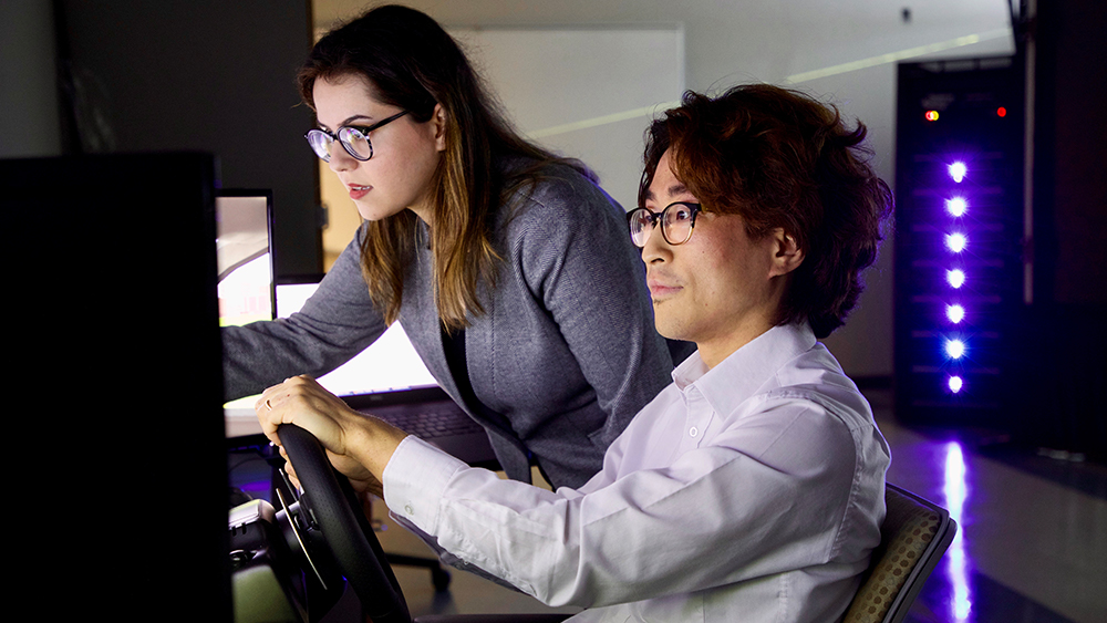 Dr. Maryam Zahabi, a woman with long brown hair and round glasses, stands and points to a computer screen. A man with wavy black hair and round glasses, wearing a white lab coat, sits beside her and watches.