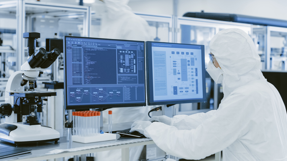 Scientist in white protective gear working on a personal computer in a modern manufacturing lab producing semiconductors and pharmaceutical items.