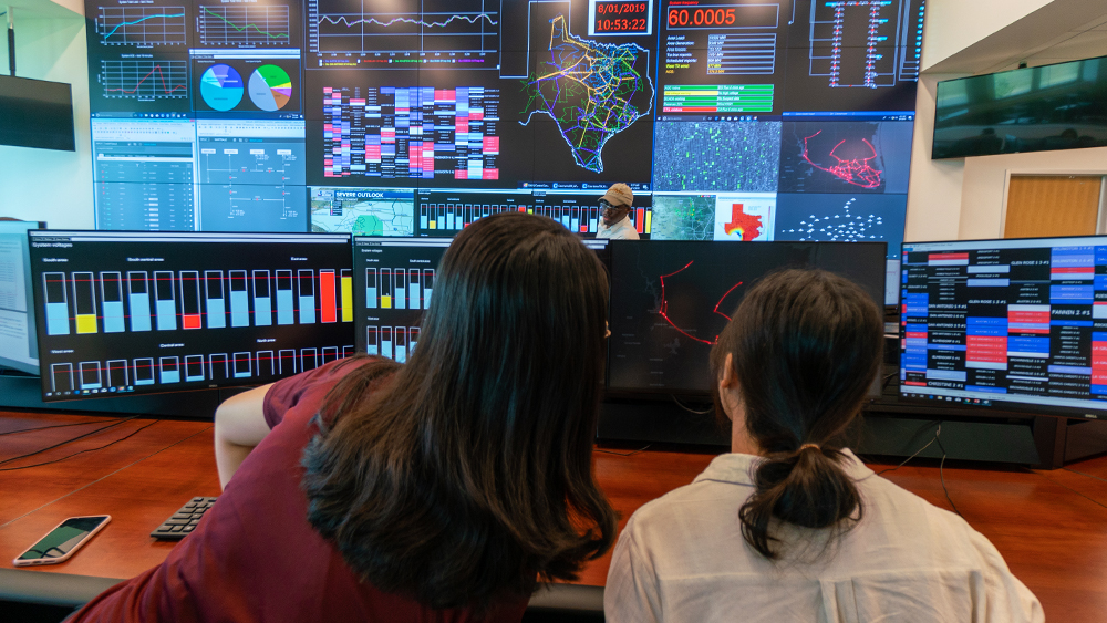 Two women with black hair sit with their backs to the camera as they view a computer screen. A wall of computer screens with a Texas maps and several numbers and graphs is on the wall ahead of them.