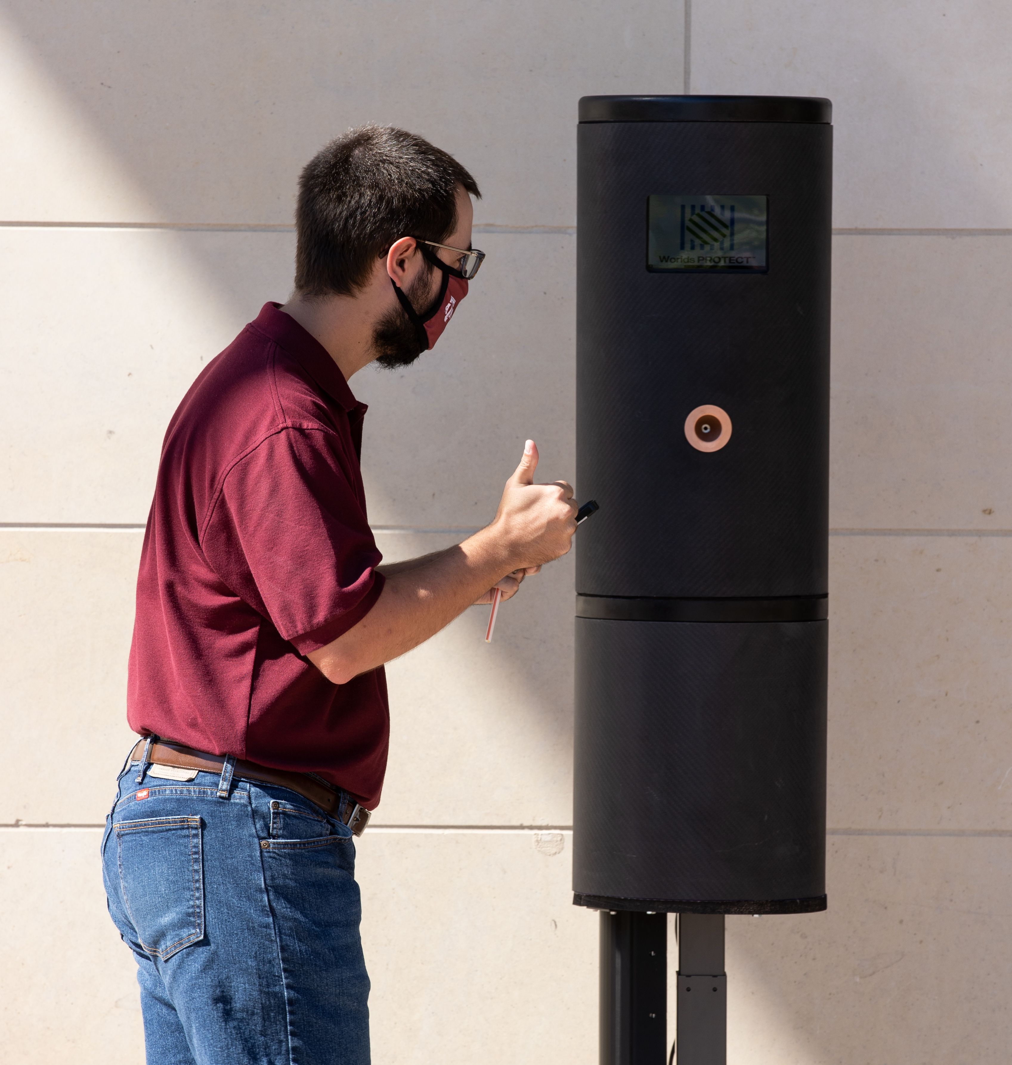 A man wearing glasses, a face mask, a maroon shirt and jeans inspects a new COVID-19 breathalyzer kiosk.