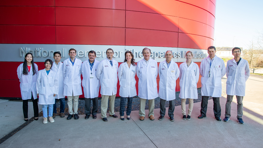 Group of 12 people in white lab coats standing in a line before the National Center for Therapeutics Manufacturing building.