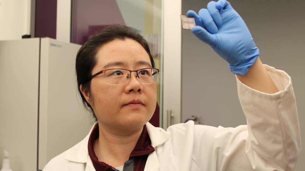 Female faculty member holds up a small square with a sensor placed on it. She's wearing glasses and blue latex gloves.
