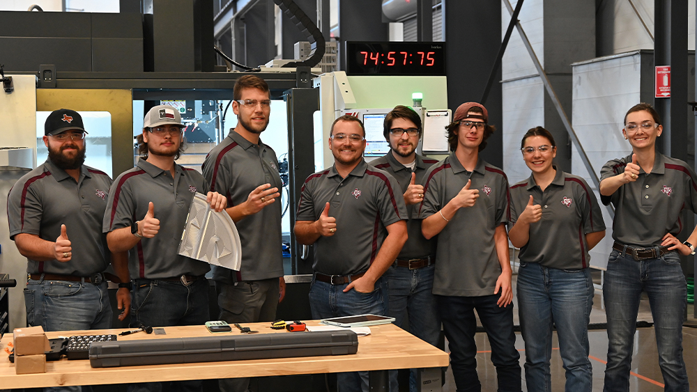 Tools sit atop a table in front of the Texas A&amp;M University team giving a thumbs up at the camera. In the background, a timer reads 74:57:75 amidst other machinery. Colton Lee holds up the team’s SEC quadrant.