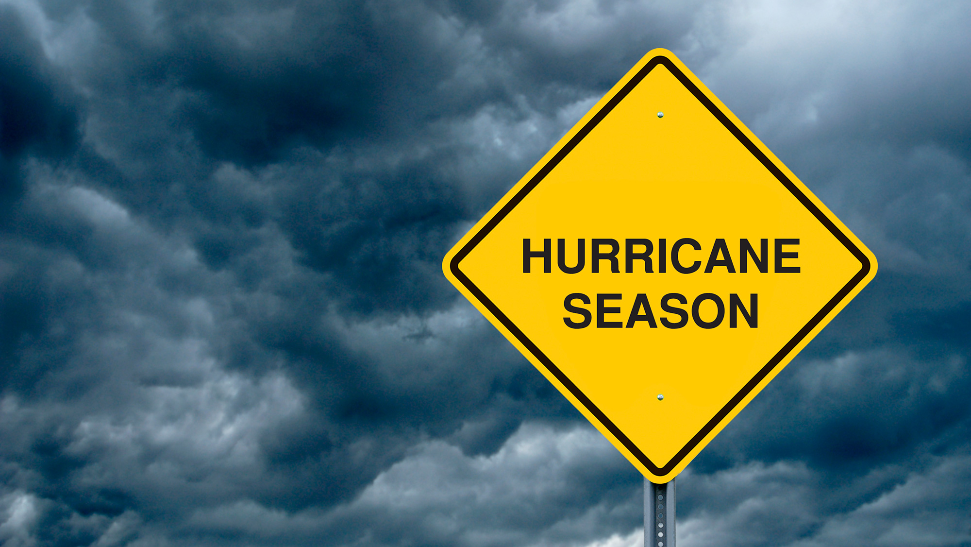 A yellow sign with black text that reads “Hurricane Season” in front of storm clouds.