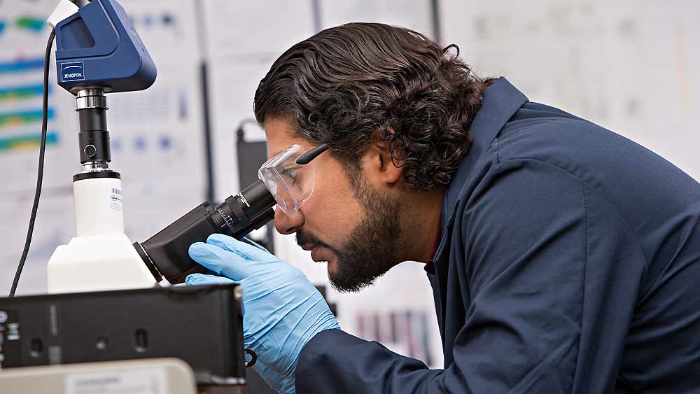 Man wearing goggles and gloves looking into a microscope.