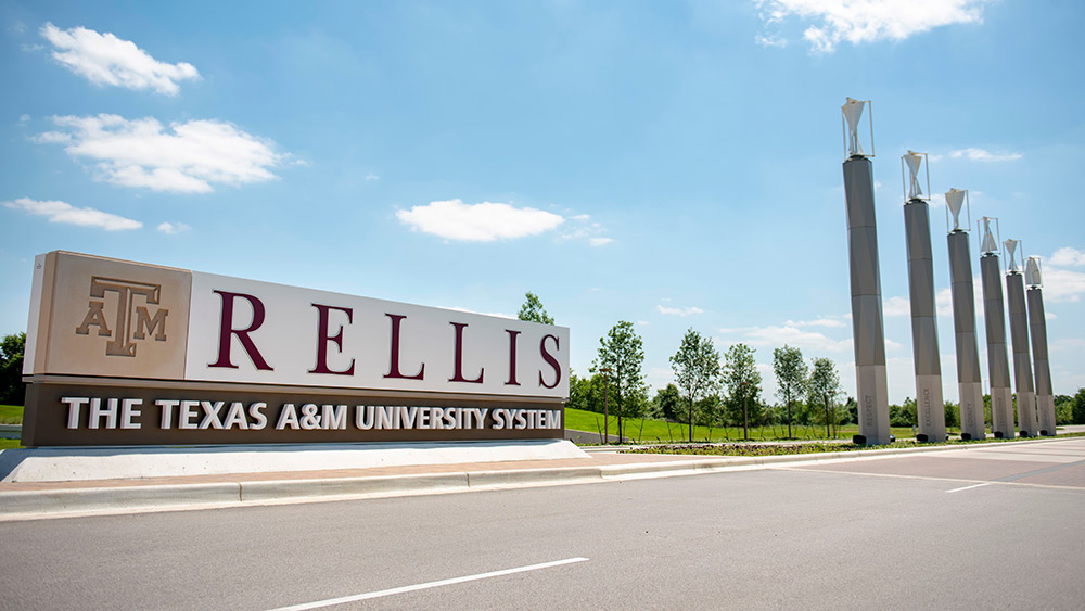 View of the entrance to RELLIS campus. There is a sign on the side of a road with the Texas A&amp;M logo and the name RELLIS, on the bottom has the name of The Texas AM University System