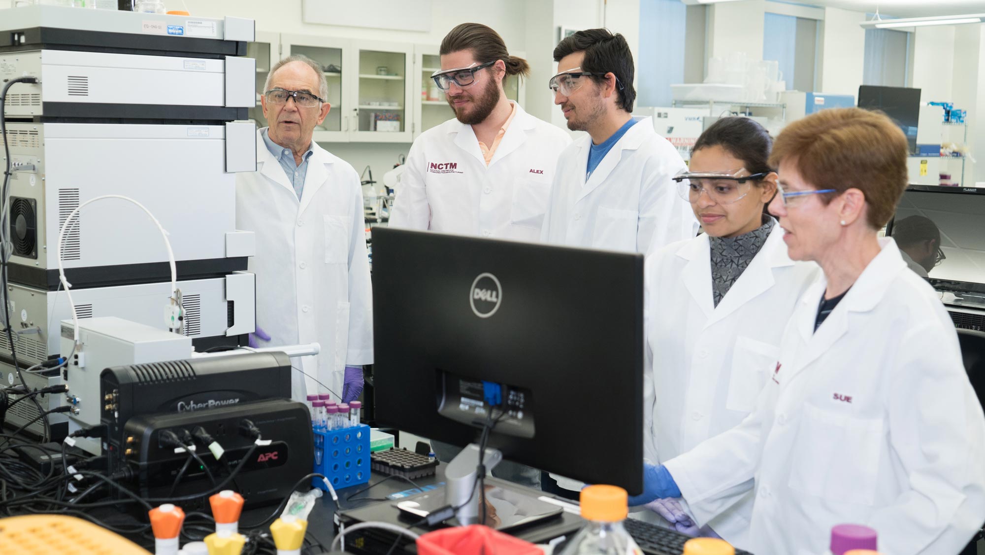 Five researchers in a lab, three male and two female, observe lab equipment and a computer monitor. All are wearing safety goggles and white lab coats.