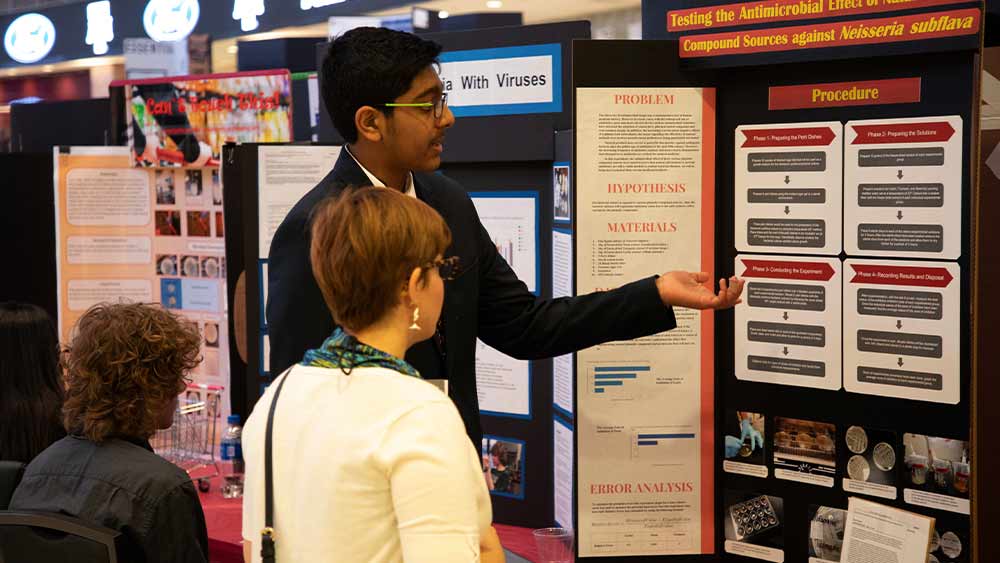 A male student pointing to and discussing his poster project with a passer-by at a past Texas Science and Engineering Fair.