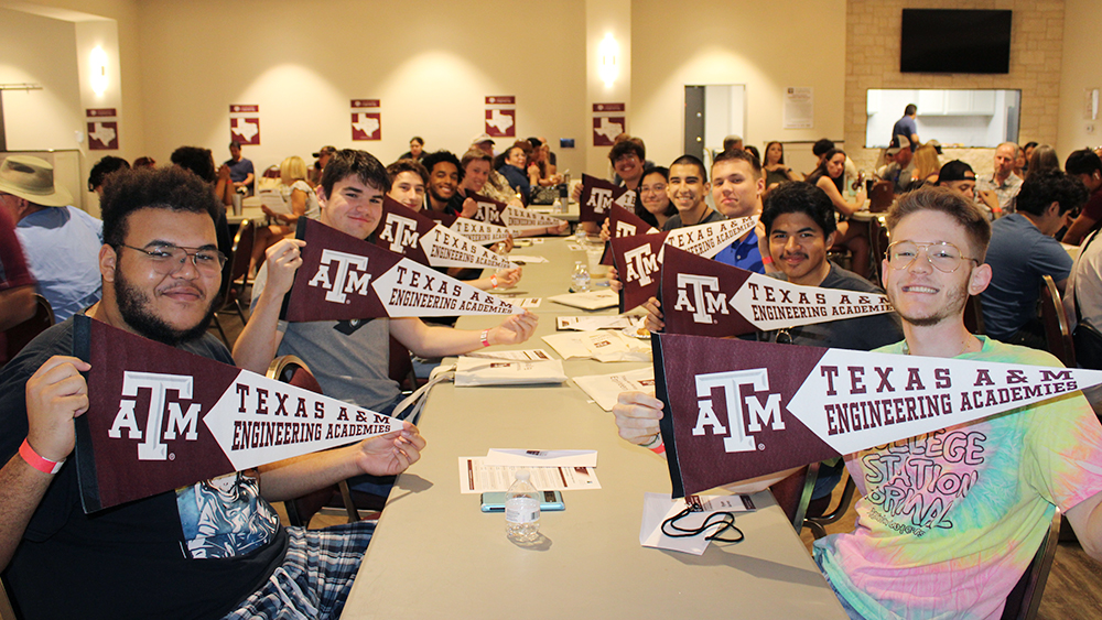 Students sit in two lines along a long rectangle table holding up pennants that say Texas A&amp;M Engineering Academies. 