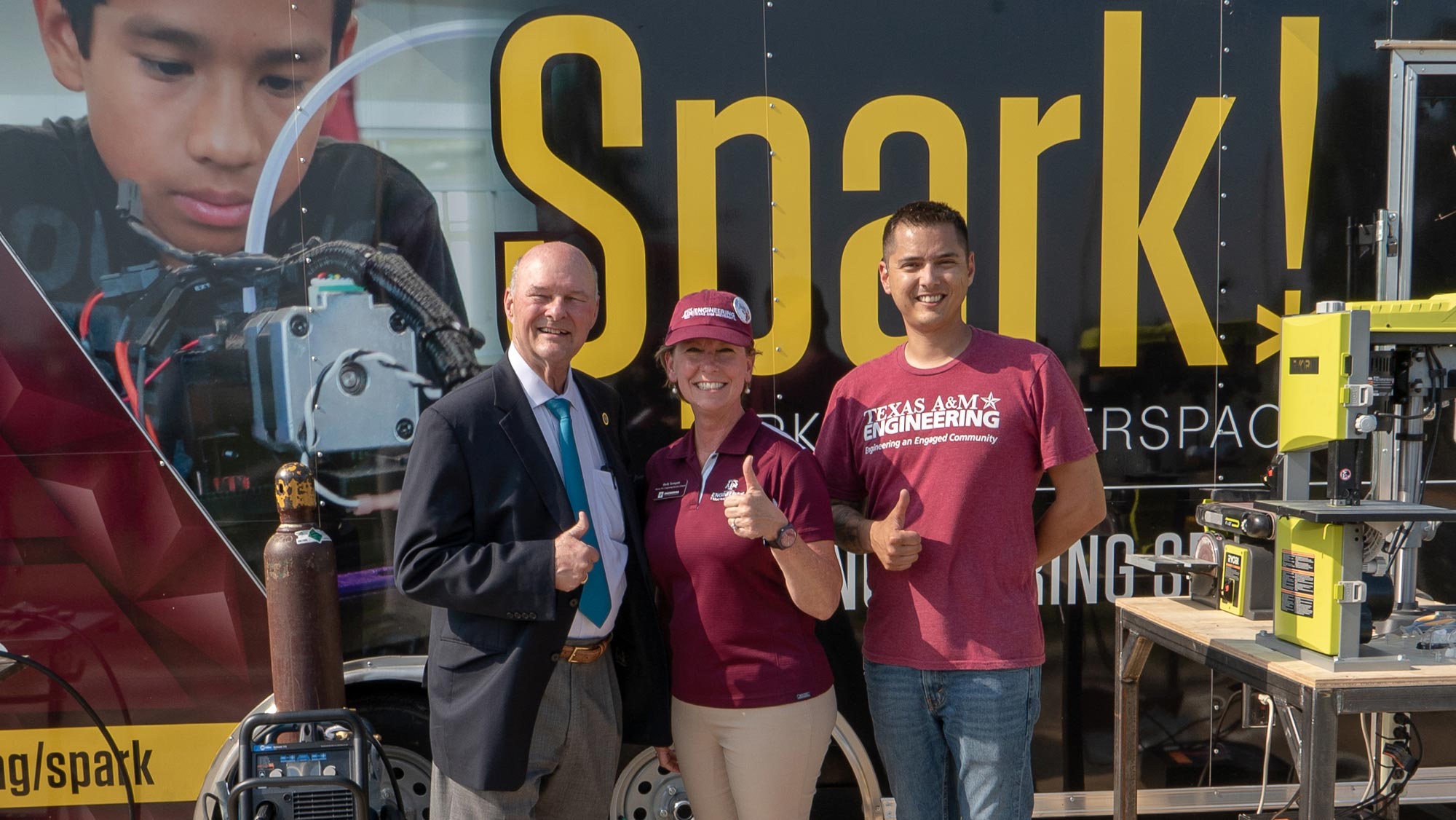 Two adult males and an adult female smile at the camera and give the "gig 'em" thumbs up symbol in front of the Spark trailer.