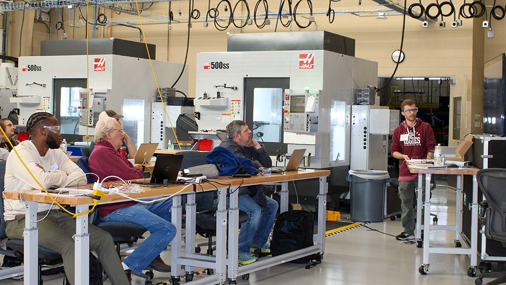 Adults sitting at desks in a warehouse setting look at a speaker standing next to a TV screen discussing the project. 