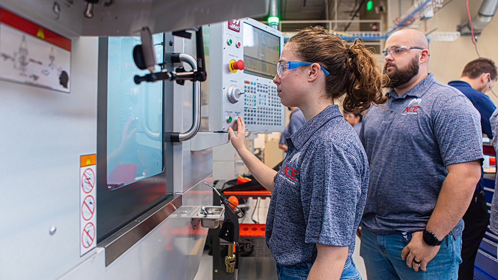 Female wearing a blue shirt uses controls on a CNC machine while man oversees her work. 