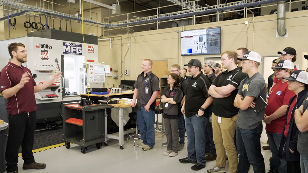 Male in maroon polo and black pants faces and talks to a group of people in a machining shop in front of a CNC machine. 