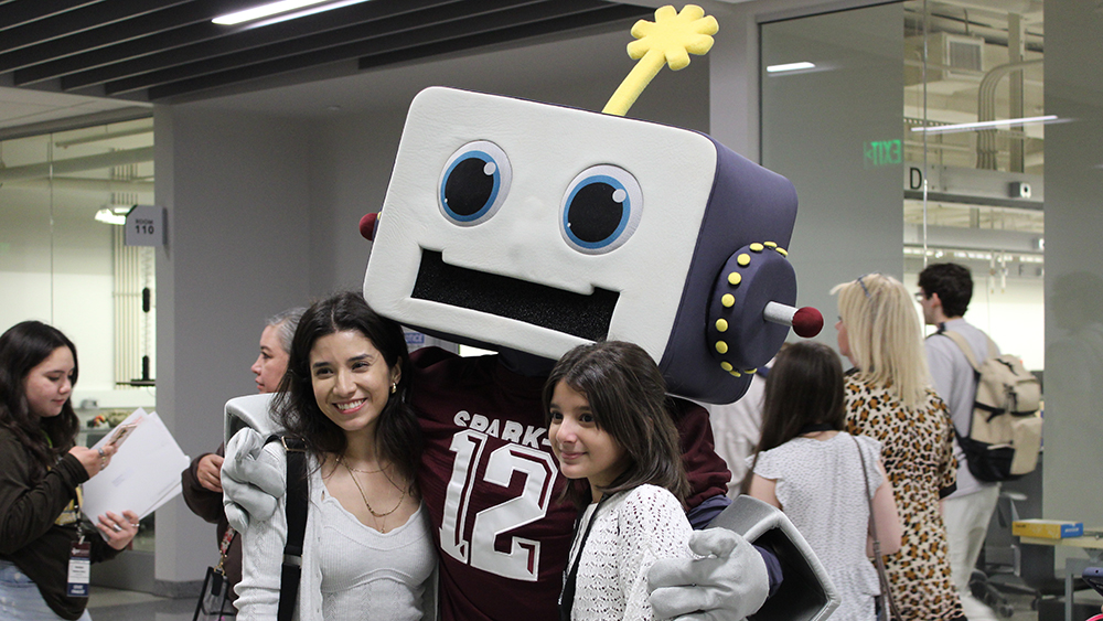 Person in robot suit and maroon football jersey wraps their arms around two young students smiling at the camera. 