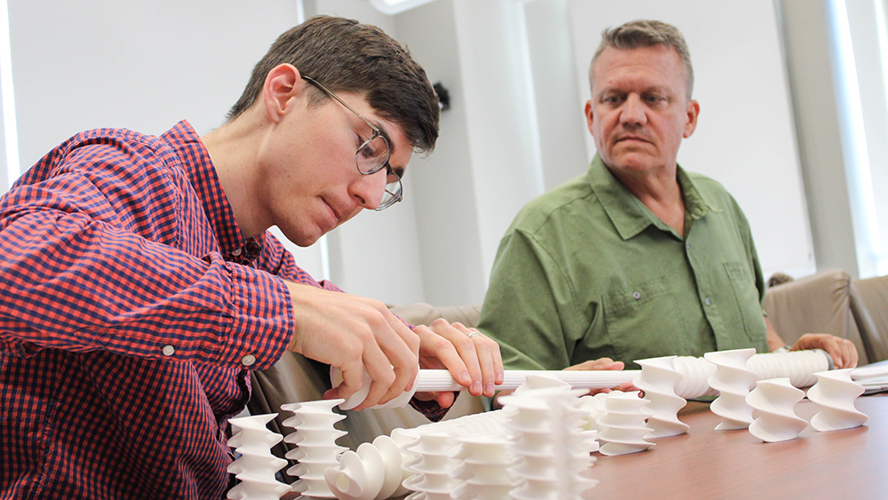 A man wearing glasses and a blue and red checkered shirt puts together white screws and tubing while another man in a green shirt looks upon. 