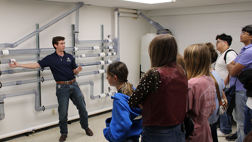 Person pointing to a series of pipes on a wall speaks to a group of high school students.  